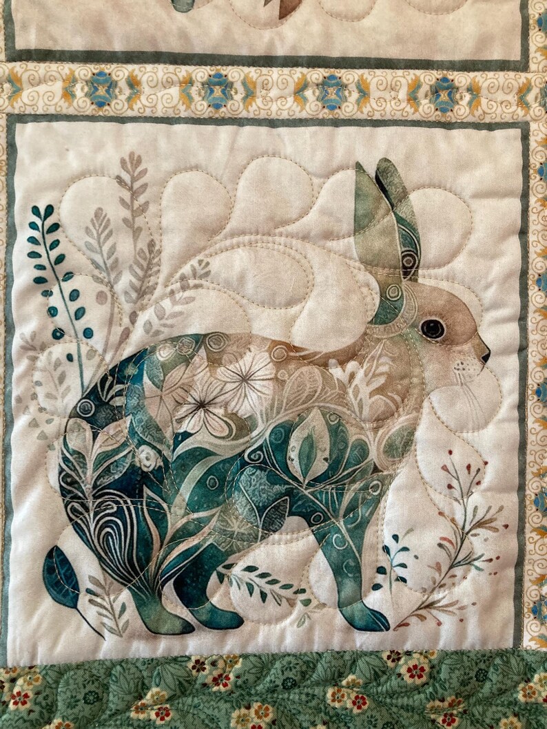 以下が含まれることがあります： ウサギの詳細なイラストが描かれた、キルト加工された布の正方形。ウサギは、ティールと白の花柄で埋められています。背景は白で、ティールと金のフローラルデザインの装飾的なボーダーが付いています。キルトはティール、茶色、白のミックスです。