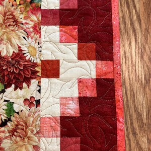 以下が含まれることがあります： 左側に花柄、右側に赤とクリーム色のパッチワーク模様が施されたキルトのテーブルランナー。 花柄の部分には、ダリアやその他のオレンジ、赤、クリーム色の花が描かれています。