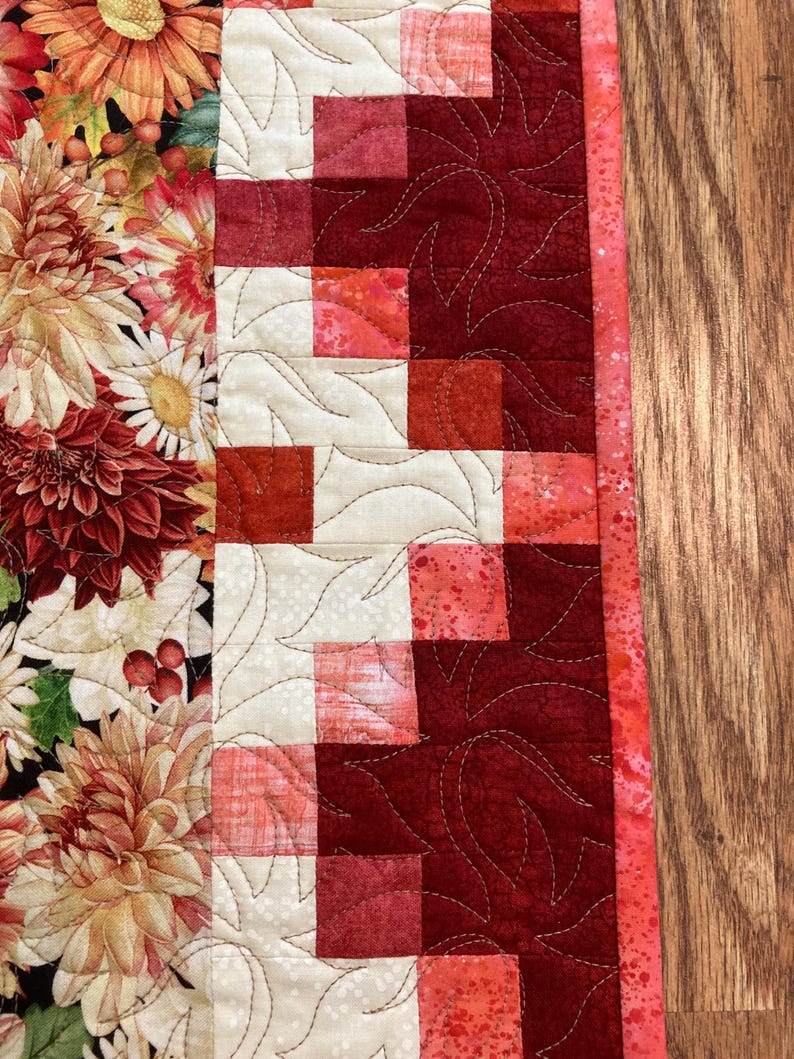 以下が含まれることがあります： 左側に花柄、右側に赤とクリーム色のパッチワーク模様が施されたキルトのテーブルランナー。 花柄の部分には、ダリアやその他のオレンジ、赤、クリーム色の花が描かれています。