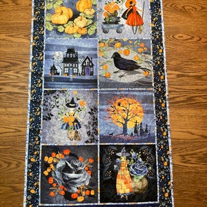 May include: A rectangular fabric table runner featuring eight distinct panels with Halloween-themed illustrations including pumpkins, a haunted house, a black crow, witches, and floral motifs, all bordered by a dark patterned fabric with orange accents.