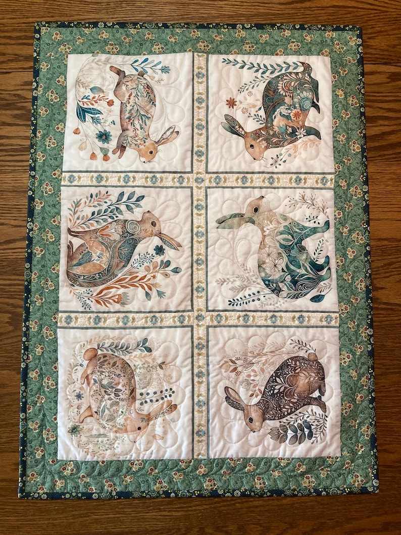 以下が含まれることがあります： 6つの正方形が特徴の装飾的なキルトテーブルランナー。それぞれに異なるウサギのイラストが描かれています。ウサギはさまざまなポーズをとっており、花や植物の要素に囲まれています。ボーダーは緑色で、花柄です。