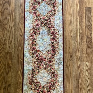 May include: A rectangular floral quilt with a burgundy border. The quilt features a repeating pattern of stylized floral shapes in shades of pink, red, and cream against a light blue background. The quilt is placed on a wooden surface.