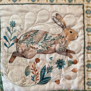以下が含まれることがあります： 茶色、ティール、オレンジ色の植物の詳細が施された、走っているウサギのイラストが描かれたキルト正方形。 ウサギは、スカラップ模様の白い背景に配置され、花のボーダーで縁取られています。