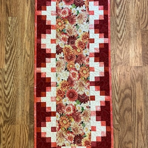 May include: A rectangular quilted table runner with a floral pattern in the center. The center features a variety of flowers in shades of red, orange, yellow, and white. The border is a dark red with a geometric pattern. The runner is approximately 40 inches long.