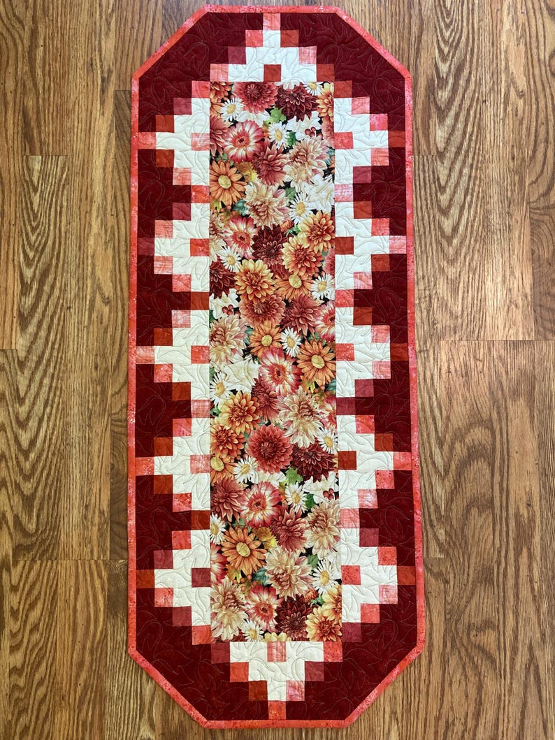 以下が含まれることがあります： 中央に花柄のある長方形のキルトテーブルランナー。中央には、赤、オレンジ、黄色、白の色合いの花が咲き乱れています。縁は幾何学模様の濃い赤色です。ランナーの長さは約102cmです。