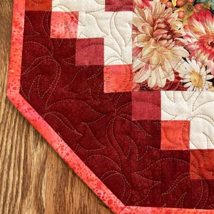 以下が含まれることがあります： 赤、クリーム、コーラルの色合いのパッチワークデザインが特徴のキルトテーブルランナー。 ランナーは片面に花柄、もう片面に葉柄があります。 エッジはコーラル色のバインディングで仕上げられています。