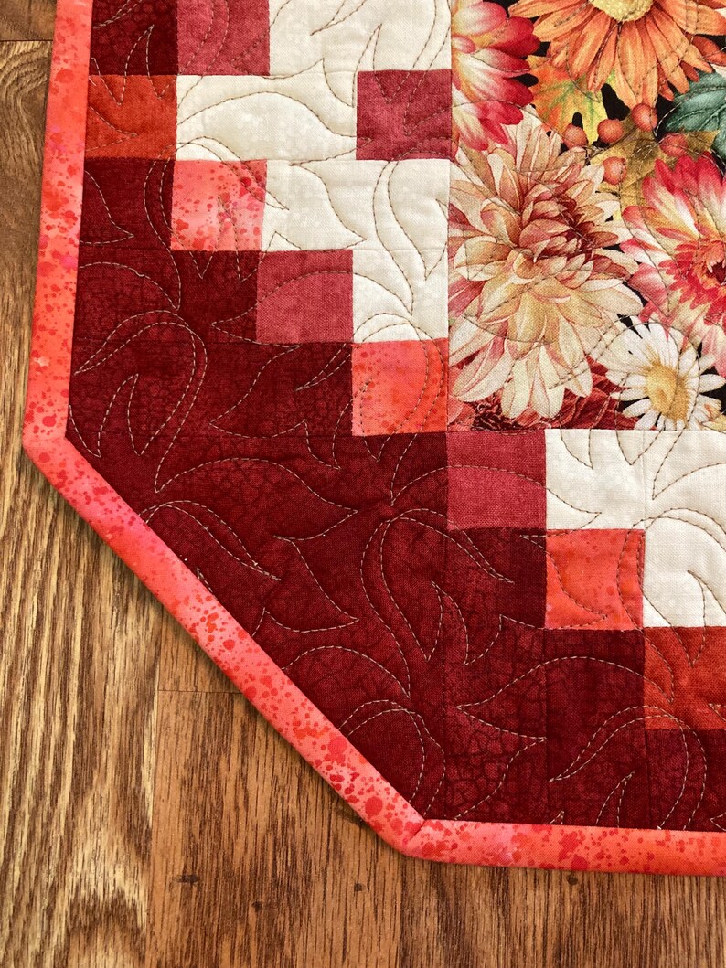 以下が含まれることがあります： 赤、クリーム、コーラルの色合いのパッチワークデザインが特徴のキルトテーブルランナー。 ランナーは片面に花柄、もう片面に葉柄があります。 エッジはコーラル色のバインディングで仕上げられています。