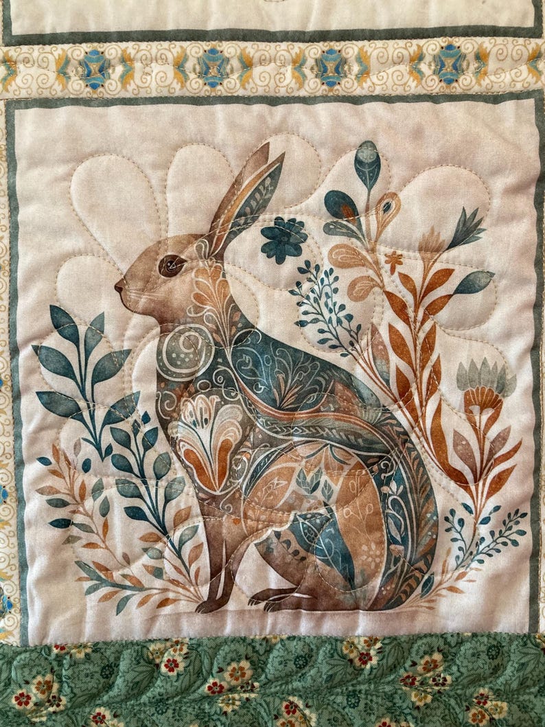 以下が含まれることがあります： 茶色、ティール、ベージュの色合いで、スタイリッシュな花の要素に囲まれたウサギの詳細なイラストが特徴のキルト壁掛け。ウサギには複雑な模様があります。ボーダーは緑とクリーム色の花柄です。