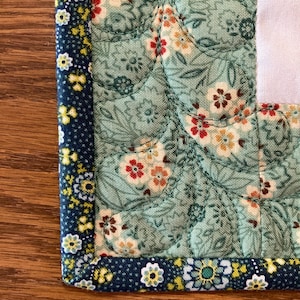 以下が含まれることがあります： キルト生地の角のクローズアップ。メインの生地は、赤、黄、白の花柄の淡いティールです。小さな黄色と白の花が付いた濃い青色のボーダーが端を縁取っています。キルティングはテクスチャ効果を生み出します。