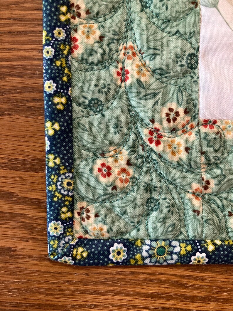 以下が含まれることがあります： キルト生地の角のクローズアップ。メインの生地は、赤、黄、白の花柄の淡いティールです。小さな黄色と白の花が付いた濃い青色のボーダーが端を縁取っています。キルティングはテクスチャ効果を生み出します。