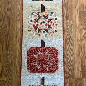 May include: A quilted table runner featuring four pumpkin designs in autumnal colors. Each pumpkin has a brown stem and is filled with fall-themed patterns, including leaves and stars. The runner has a red border.