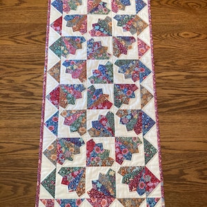 May include: A white table runner with a floral pattern. The runner is made up of squares with a unique design in each square. The squares are surrounded by a pink border.