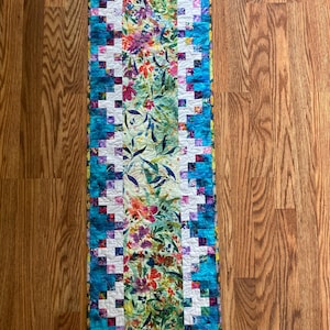 May include: A rectangular quilted table runner with a floral design. The center features a vibrant floral pattern with red, purple, and green flowers. The border is a patchwork of blue and white squares, with a teal edge.