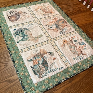 以下が含まれることがあります： 6つのパネルにウサギのイラストが描かれたキルトのテーブルランナー。各ウサギは、茶色、ティール、クリーム色の花柄と植物柄で描かれています。縁は緑色で、花柄です。