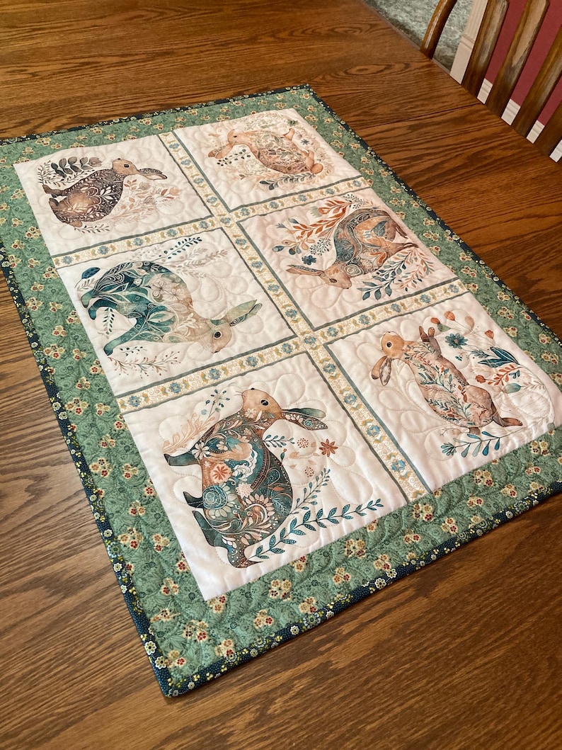 以下が含まれることがあります： 6つのパネルにウサギのイラストが描かれたキルトのテーブルランナー。各ウサギは、茶色、ティール、クリーム色の花柄と植物柄で描かれています。縁は緑色で、花柄です。