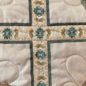 以下が含まれることがあります： 十字のデザインが施されたキルト生地のクローズアップ。十字は、薄緑色のボーダーで形成され、青と金のフローラルモチーフが特徴の装飾リボンで装飾されています。背景は、キルト模様の薄いクリーム色です。