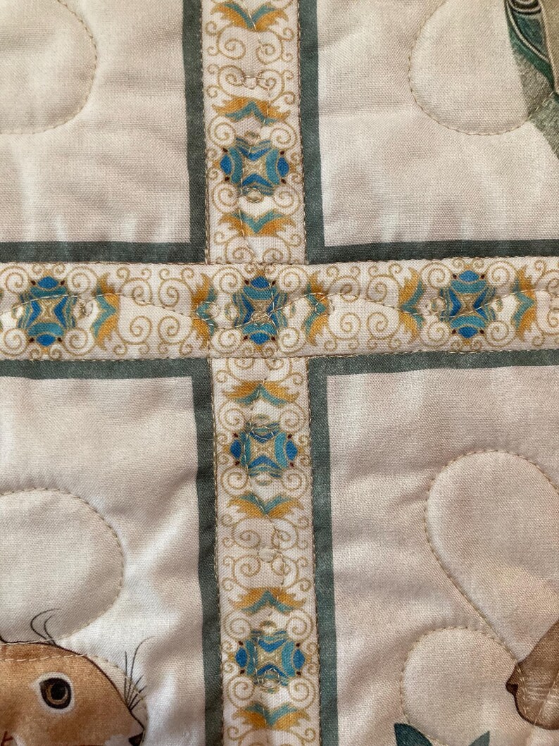 以下が含まれることがあります： 十字のデザインが施されたキルト生地のクローズアップ。十字は、薄緑色のボーダーで形成され、青と金のフローラルモチーフが特徴の装飾リボンで装飾されています。背景は、キルト模様の薄いクリーム色です。