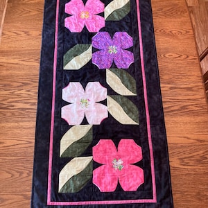May include: A rectangular quilted table runner featuring a black background with a pink border. The design includes four stylized flowers in shades of pink and purple, interspersed with several light and dark green fabric leaves.