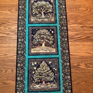 May include: A rectangular quilted table runner featuring three square panels, each depicting a different tree scene against a dark background. The panels are framed by a bright teal border and an outer border with a dense floral pattern.