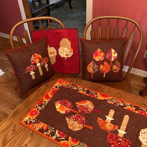 May include: A brown table runner with a fall leaf design in shades of orange, red, and brown. The runner is surrounded by a border of sunflowers and leaves. There are three pillows on the chairs behind the table runner, two with a mushroom design and one with a leaf design.