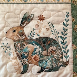 以下が含まれることがあります： ウサギのデザインが施された装飾的なキルト生地パネル。ウサギは、ティール、ブラウン、クリームの色合いの花柄とペイズリー柄で装飾されています。背景は白で、装飾的なボーダーが付いています。