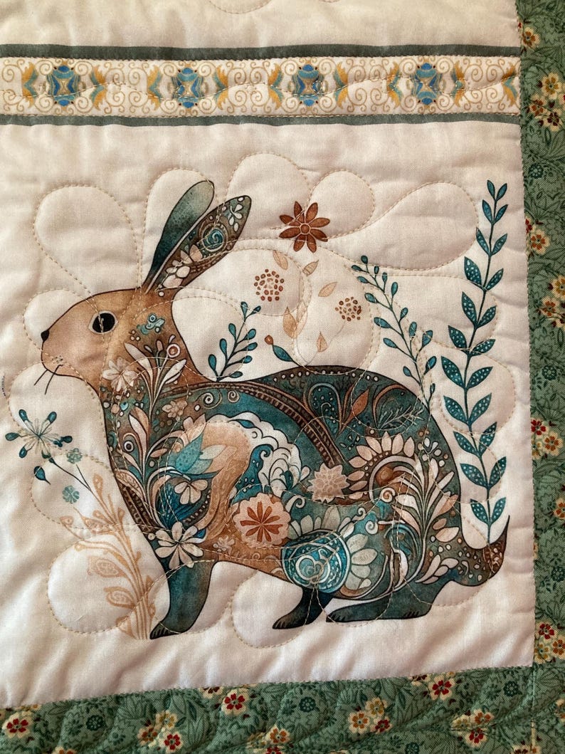 以下が含まれることがあります： ウサギのデザインが施された装飾的なキルト生地パネル。ウサギは、ティール、ブラウン、クリームの色合いの花柄とペイズリー柄で装飾されています。背景は白で、装飾的なボーダーが付いています。