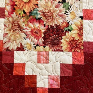 以下が含まれることがあります： 黒い背景にオレンジ、赤、黄色のダリアの花柄が特徴のキルトテーブルランナー。縁は赤と白の色合いのパッチワーク模様で、葉の形をしたキルティングが施されています。