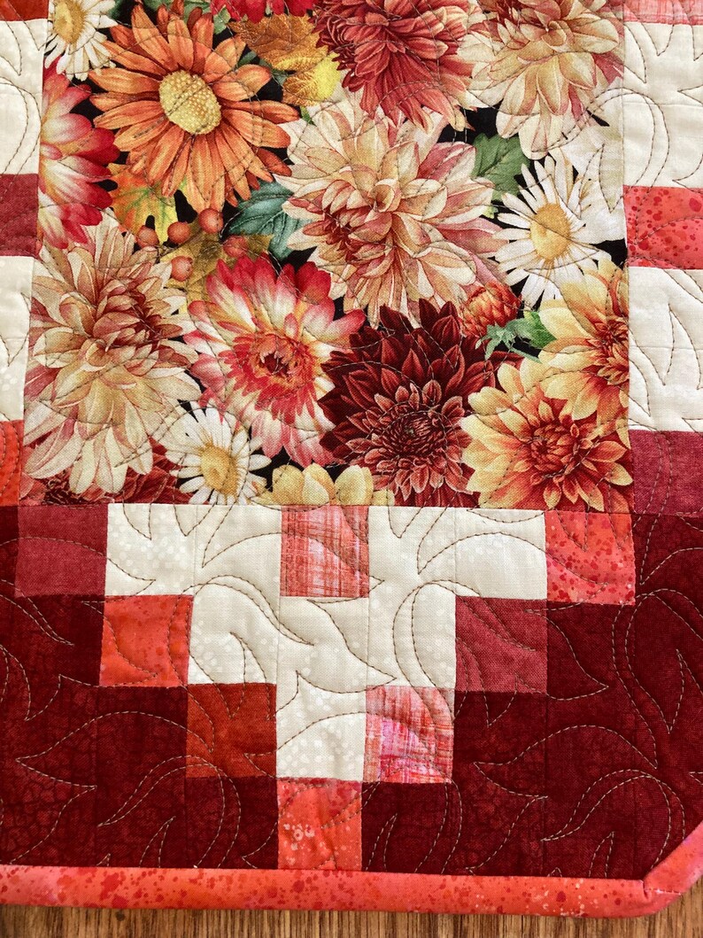 以下が含まれることがあります： 黒い背景にオレンジ、赤、黄色のダリアの花柄が特徴のキルトテーブルランナー。縁は赤と白の色合いのパッチワーク模様で、葉の形をしたキルティングが施されています。