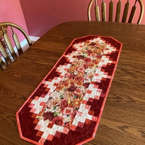 以下が含まれることがあります： 花柄のデザインが施されたキルトのテーブルランナー。中央には、赤、オレンジ、黄色の鮮やかな花が配置されています。ランナーは、赤と白のブロックと赤い縁取りで幾何学模様になっています。