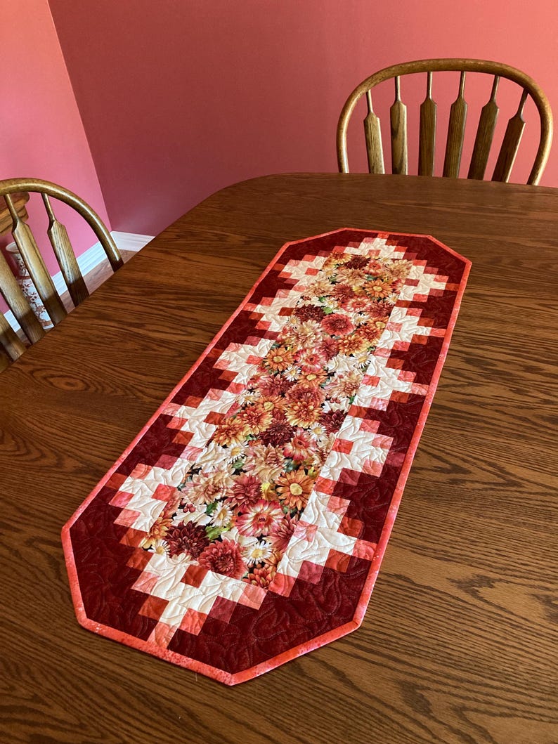 以下が含まれることがあります： 花柄のデザインが施されたキルトのテーブルランナー。中央には、赤、オレンジ、黄色の鮮やかな花が配置されています。ランナーは、赤と白のブロックと赤い縁取りで幾何学模様になっています。