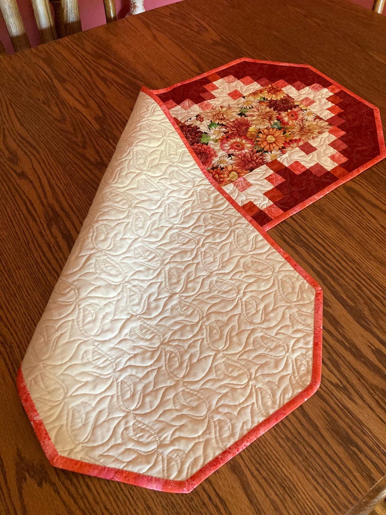 以下が含まれることがあります： 花柄と赤い縁取りのキルトテーブルランナー。ランナーは、キルト模様のクリーム色の裏地が特徴です。花柄のデザインには、赤、オレンジ、黄色の色合いが含まれています。ランナーは木製のテーブルの上にあります。