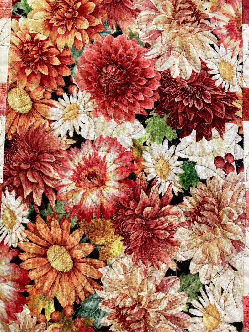 以下が含まれることがあります： 赤、オレンジ、クリーム色のダリアとデイジーが黒い背景に描かれた、花のキルトのクローズアップ。キルトは白い糸で縫い付けられており、テクスチャ効果を生み出しています。テキスタイルアートの美しい例です。