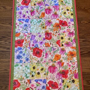May include: A rectangular quilted table runner featuring a patchwork design of various floral patterns in shades of red, orange, yellow, pink, purple, and blue. The quilt is bordered by a wide olive green fabric and a thin coral red outer edge.