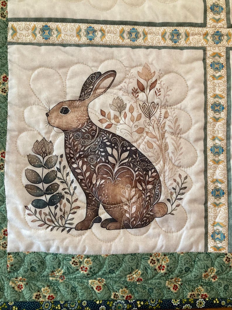 以下が含まれることがあります： 茶色のウサギと花柄のイラストが施されたキルトの壁掛け。 ウサギは、茶色と緑の色合いの様式化された植物に囲まれ、スカラップ模様のクリーム色の背景に配置されています。 縁は花柄の緑色です。