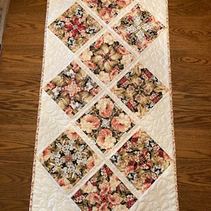 May include: A white quilted table runner with a floral pattern. The runner is made up of nine squares, each with a different floral print. The squares are arranged in a diamond pattern. The runner has a white border with a small floral print.