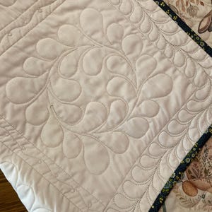 以下が含まれることがあります： クリーム色の背景のキルトテキスタイルのクローズアップ。キルトは、渦巻く涙の形をしたパターンの中心的なデザインが特徴です。エッジは花柄と濃い青色のトリムで縁取られています。