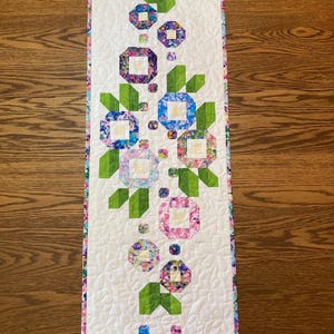 May include: A quilted table runner featuring a floral design. The runner has a white background with a repeating pattern of colorful, geometric flowers and green leaves. The edges are finished with a patterned border. The runner is approximately 12 inches wide and 36 inches long.