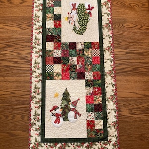 May include: A white quilted table runner with a green and red patchwork design. The runner features a snowman and Christmas tree applique in the center.