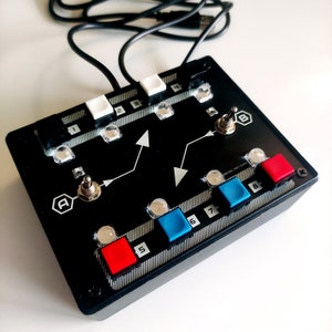 Peut inclure: Dispositif électronique noir avec huit boutons, deux interrupteurs à bascule et un câble USB. Les boutons sont numérotés de 1 à 8 et ont des dessus de couleur rouge et bleue. Les interrupteurs à bascule sont étiquetés "A" et "B".
