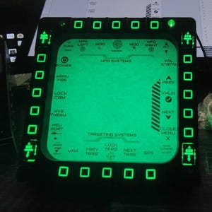 May include: A black electronic display with a green illuminated interface. The screen displays text such as "MFD SYSTEMS", "TARGETING SYSTEMS", and various function labels. Green square lights surround the screen.