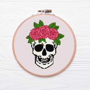 May include: A white skull with black eye sockets and a toothy grin, wearing a crown of three pink roses with green leaves.