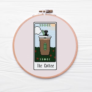Puede incluir: Una bordado de punto de cruz de una carta de tarot con una taza de café y un gato negro. La carta se titula "The Coffee".