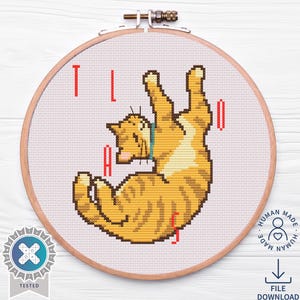 May include: Cross-stitch embroidery of a ginger cat in a playful pose, inside a wooden hoop. The word "TAIL" is stitched in red above and below the cat. The design is on a white fabric, with a "Tested" badge and a "Human Made" logo.