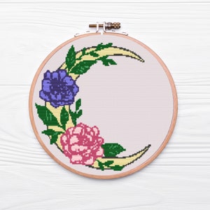 May include: A cross-stitch embroidery hoop featuring a crescent moon design. The moon is adorned with purple and pink flowers and green leaves.