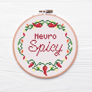 May include: A white embroidery hoop with a red and green embroidered design of chilli peppers and vines. The text "Neuro Spicy" is embroidered in red.