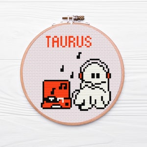May include: A white wooden embroidery hoop with a beige fabric background. The fabric has a cross-stitch design of a ghost wearing headphones and listening to a red boombox. The text "TAURUS" is stitched in orange above the design.