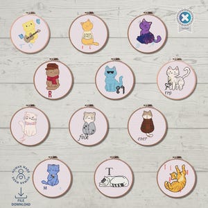 May include: A collection of twelve embroidered cat portraits in wooden hoops. Each cat is depicted in a unique pose and style, with some wearing accessories. The hoops are arranged on a light wood-grain surface. Some hoops have text, including the letters "T", "R", "M", "F", and "rep".