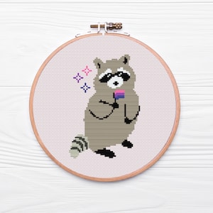 Könnte beinhalten: Kreuzstich-Stickerei eines Waschbären, der eine bisexuelle Pride-Flagge hält. Der Waschbär ist grau und weiß mit schwarzen Akzenten. Rosa und blaue Sternausbrüche befinden sich über dem Waschbären. Die Stickerei befindet sich in einem Holzreifen.