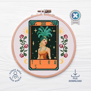 May include: A cross-stitch embroidery in a wooden hoop features a tarot card design. The card depicts a woman with a palm frond headdress. The design includes the text "TLOA" and is surrounded by floral and celestial motifs.