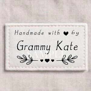 以下が含まれることがあります： 「Handmade with ♥ by Grammy Kate」の黒い文字が入った白い長方形のラベル。葉とハートの装飾的なボーダーが付いています。背景は明るい生地です。